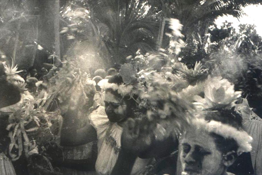 Figures adorned with elaborate floral and feathered headwear gather amidst lush tropical foliage.