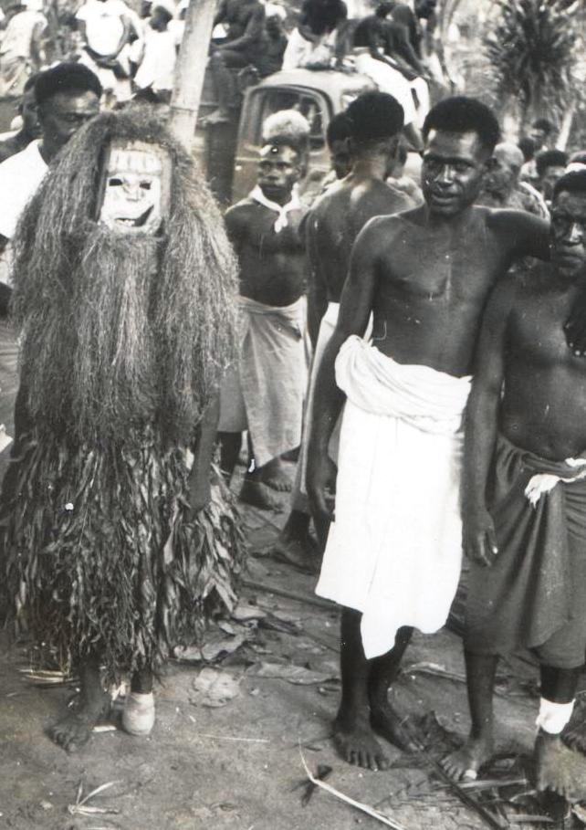 A masked performer in a grass costume stands with men at an outdoor gathering, a vehicle visible.