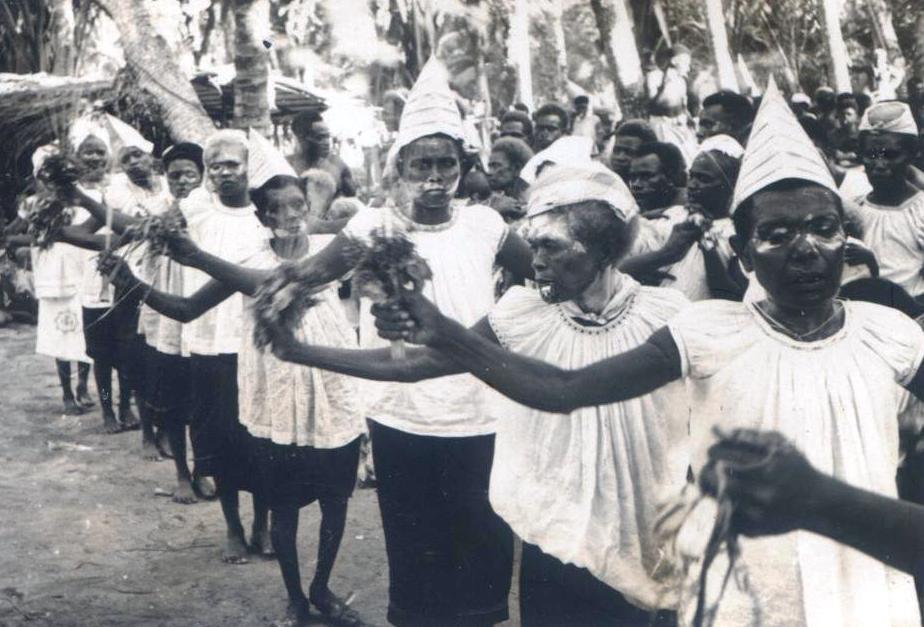 Women wearing ceremonial headwear and face paint hold foliage during a traditional procession.