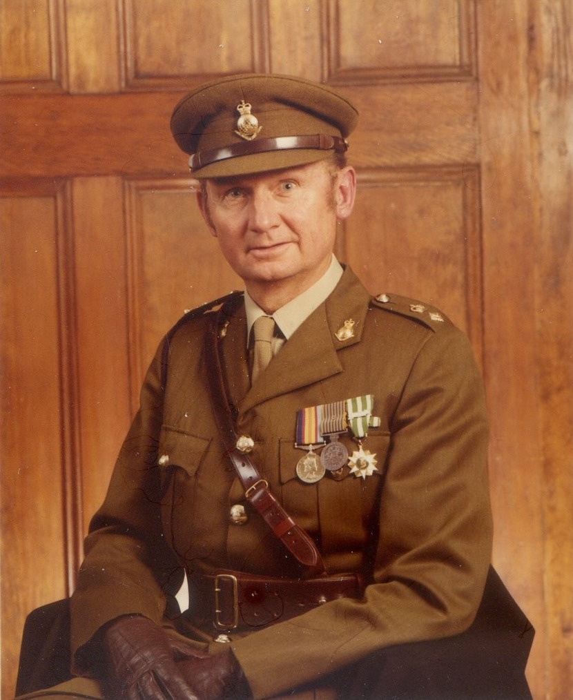 Lieutenant Colonel Bob Skitch in Army service dress and peaked cap, wearing his medals and a Sam Browne belt, seated in front of a panelled wooden door.